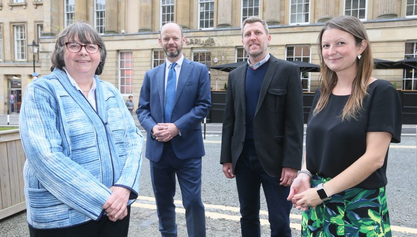 Four people stood smiling towards camera with sandstone building in background. On the left stands a woman in a light blue blazer with a short bob haircut and glasses. Next to her stands a man with no hair in a blue suit and tie and white shirt, he has a beard. Next to him stands a man with a grey blazer and blue jumper with light, cropped hair. Next to him stands a woman with longer brown hair, a black blouse and green tropical print skirt. They are all smiling towards the camera.