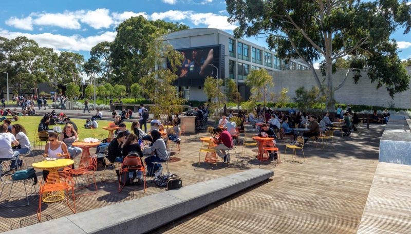Colourful outdoor seating area with tables and chairs filled with people eating, reading, studying on a campus.
