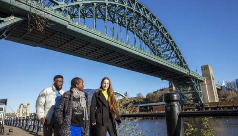 Image of three people walking by the River Tyne with the Tyne Bridge over them