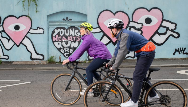 Two people cycling past graffiti on a wall