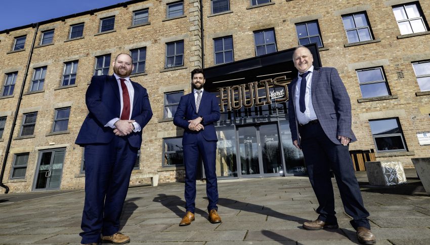 Three people in suits stood outside of Hoults Yard office building smiling to camera.