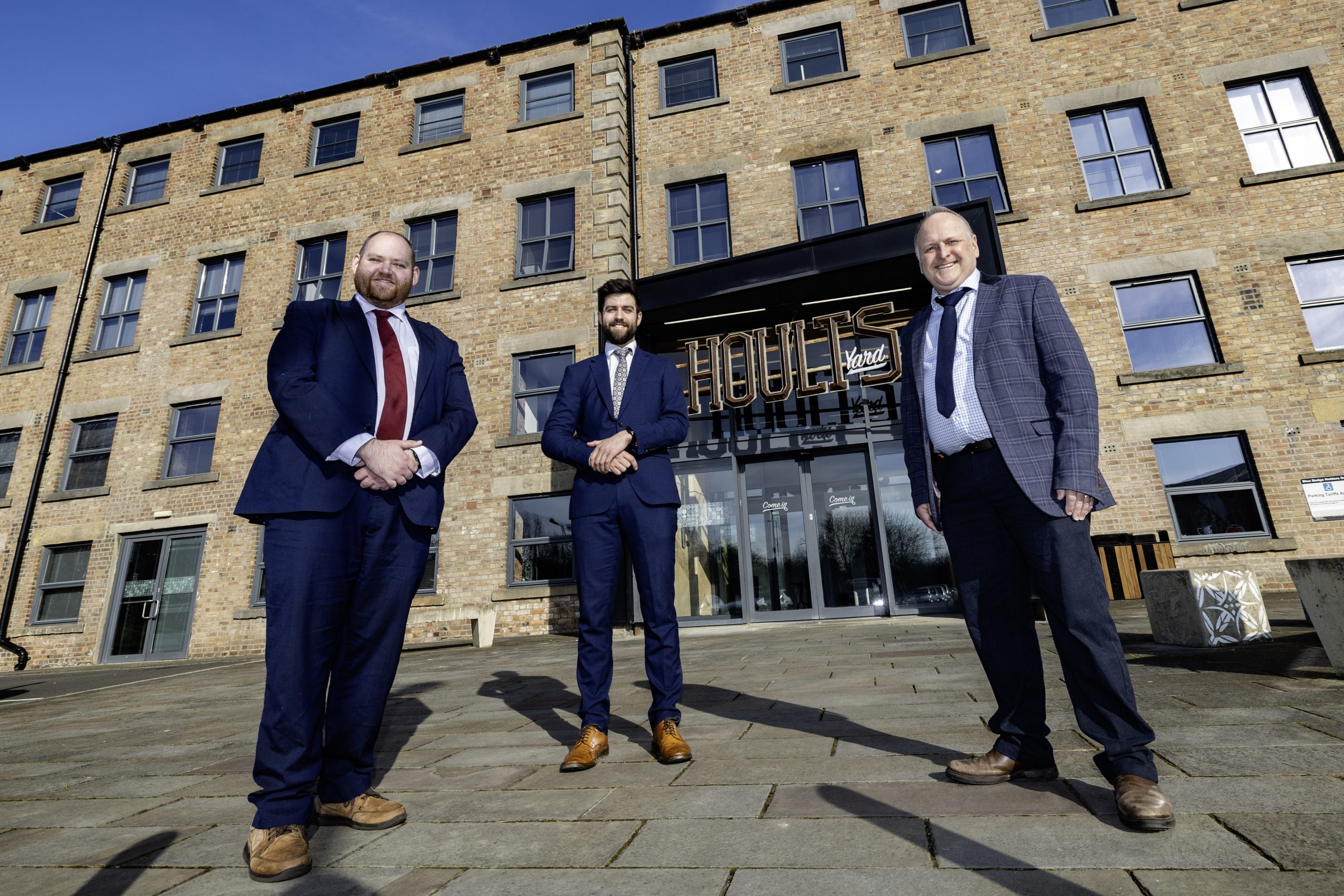 Three people in suits stood outside of Hoults Yard office building smiling to camera.