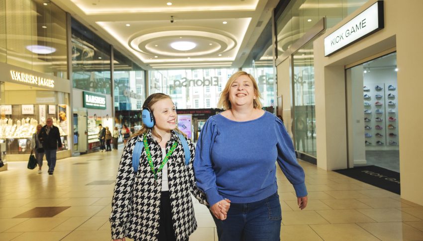 Mother and daughter walking hand-in-hand through shopping centre. Daughter is wearing ear defenders and a sunflower lanyard.