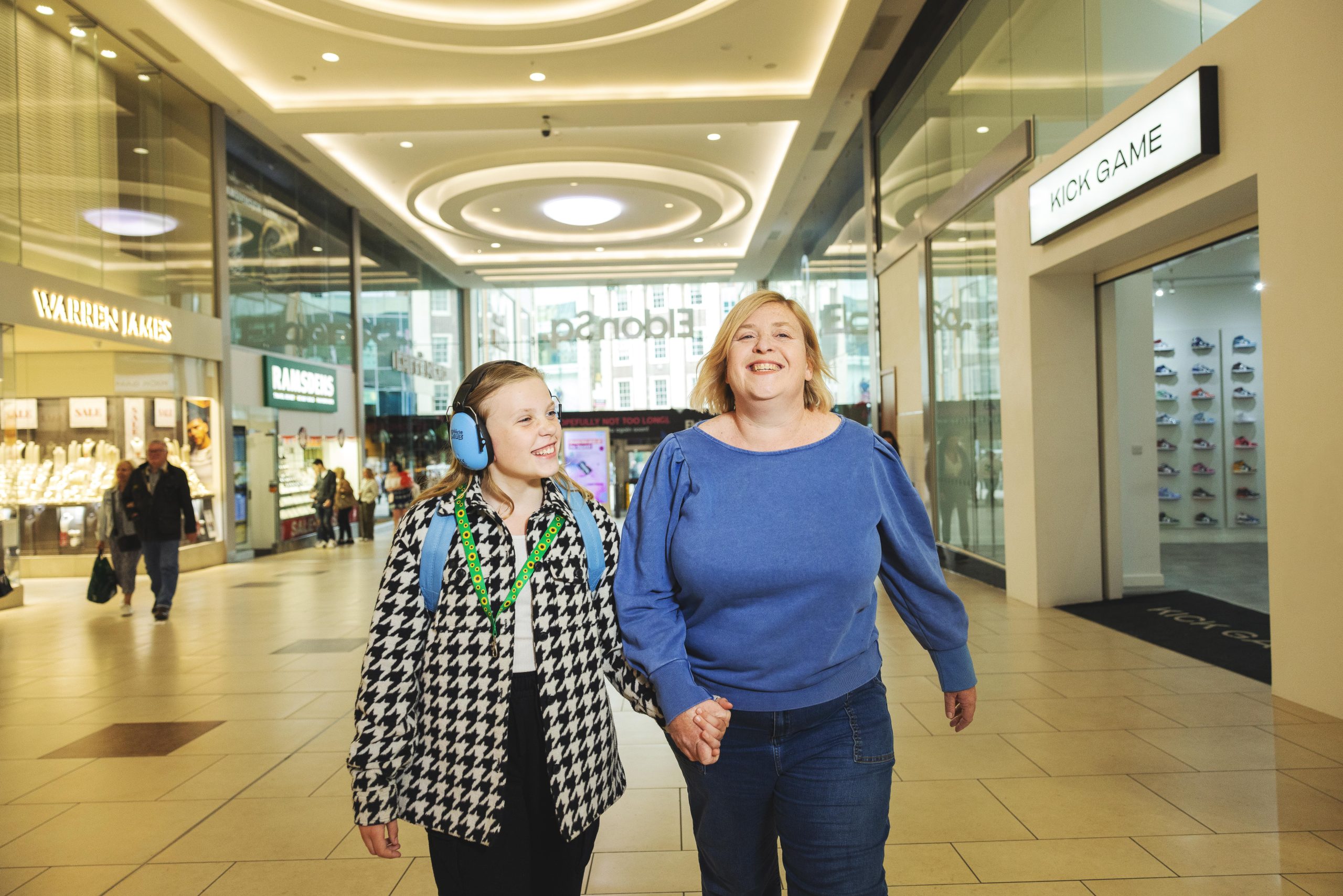 Mother and daughter walking hand-in-hand through shopping centre. Daughter is wearing ear defenders and a sunflower lanyard.