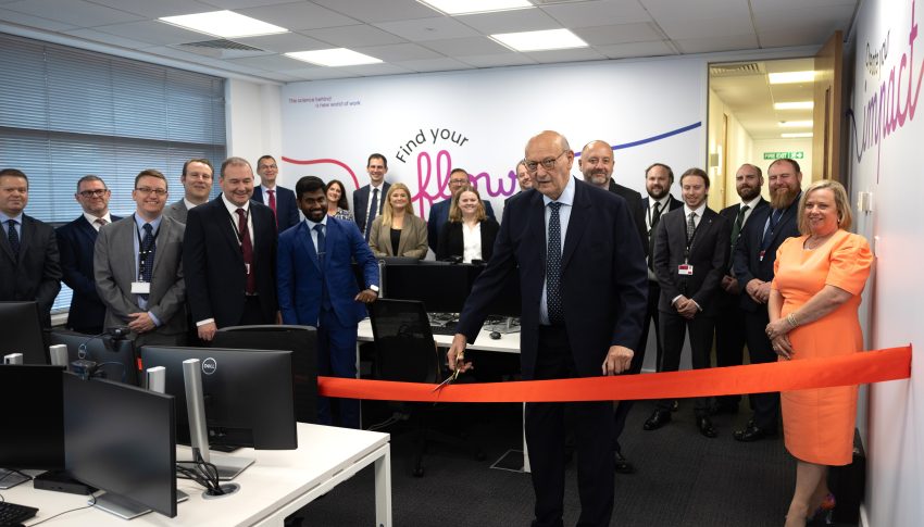 Small office room full of people and a red ribbon in the foreground being cut by a person in a suit.