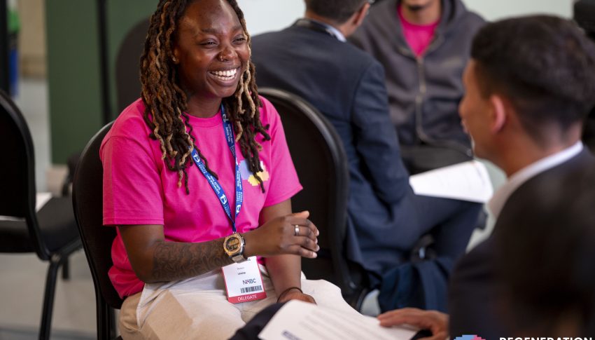 Young people smiling and talking together in a room wearing lanyards and holding sheets of paper.