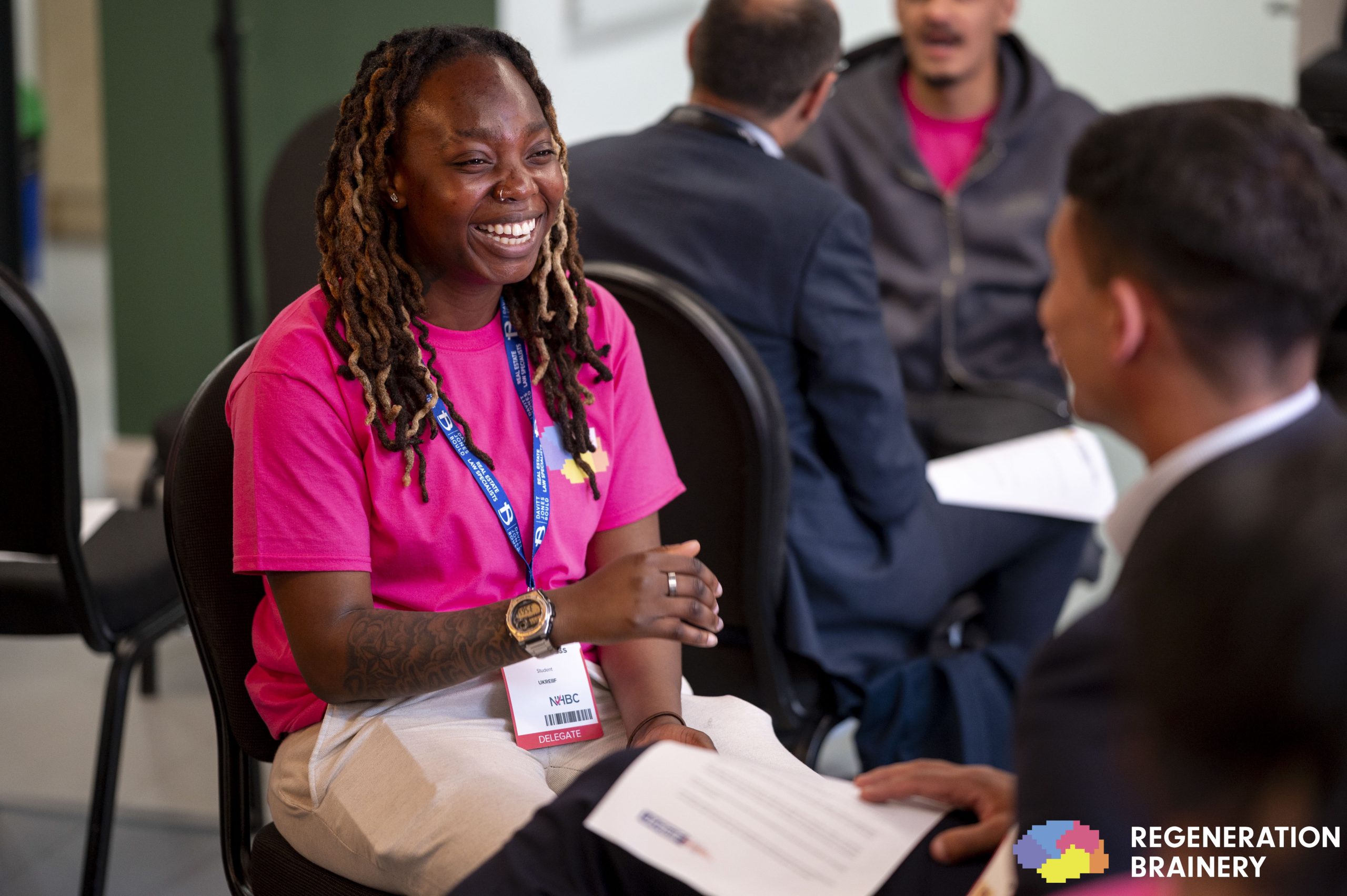Young people smiling and talking together in a room wearing lanyards and holding sheets of paper.