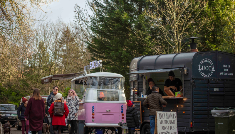 Two food trucks surrounded by trees and people and families milling around.
